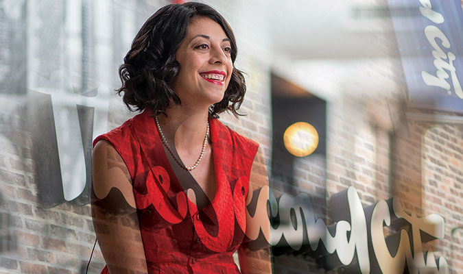 A sitting, smiling woman with red lipstick and short, dark, curly hair wearing a red dress and a white pearl necklace. Behind her is a brick wall and segments of a banner and an exterior theatre sign that read 'Second City'
