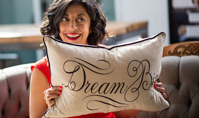 A woman shows her smiling and looking off-camera, while holding a beige couch cushion that reads 'Dream Big' in black lettering.