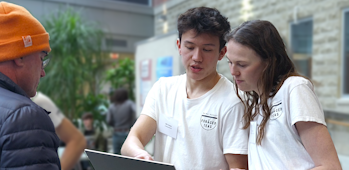 Two individuals from the Foraged Teas team at the Project SOY Plus finale engage in conversation with a member of the public. The setting is indoors, with informational posters on stands in the background.