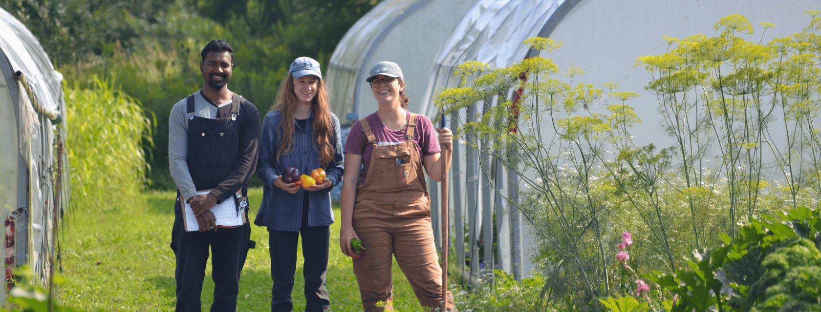 people standing outside of the organic farm, one is holding a hoe, another holding vegetables