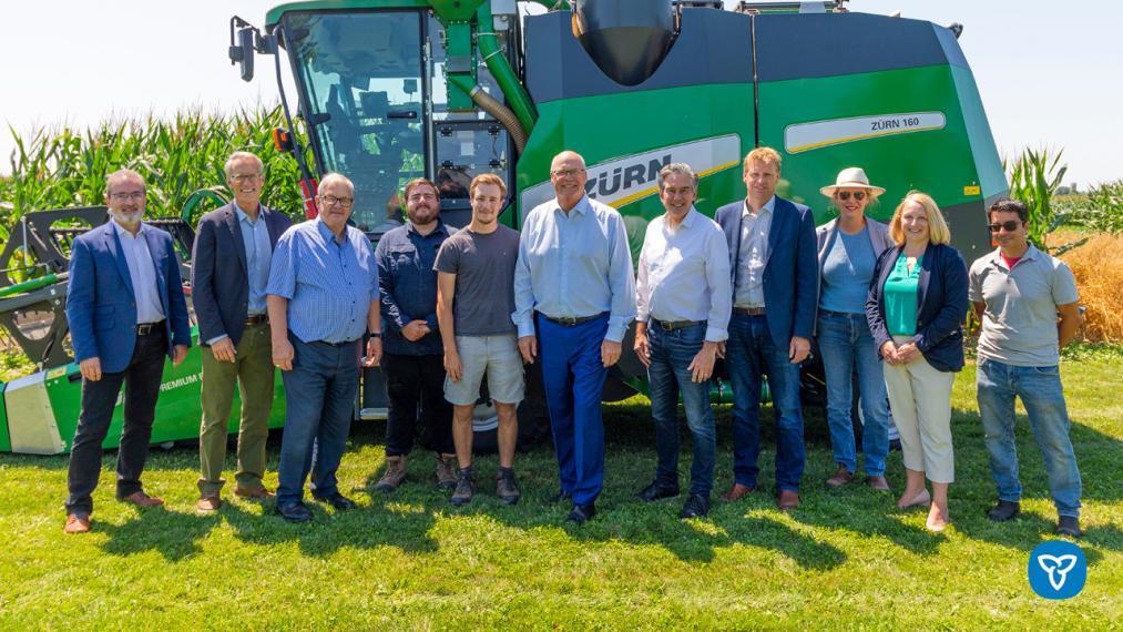 A group photo of 11 researchers and politicians stand in front of a tall green combine machine, smiling on a sunny day