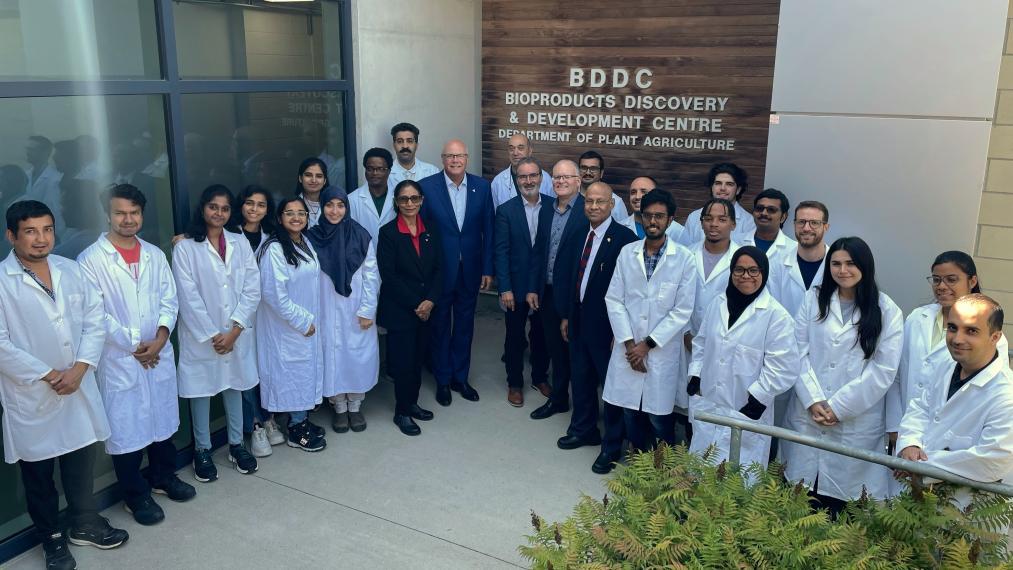 21 researchers in white lab coats stand outside a "BDDC" sign with Dr. Misra and Dr. Mohanty, Minister Flack, Dr. Van Acker and Dr. Earl, who dressed in suits.