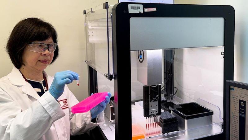 Dr. Shu Chen, a woman who wears glasses, a lab coat and gloves, peers at a sample in the lab.