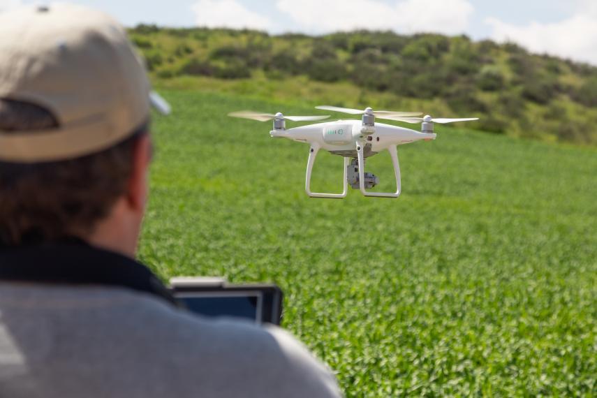 A small drone with three propellors is flying over a green farmers' field, being controlled by a farmer with a handheld device