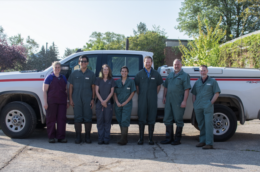 Seven veterinarians, all wearing coveralls, stand in front of a U of G truck