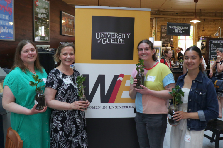 From left to right: Dr. Jana Levison, Jean Hein, Samantha Rutherford and Sofia Rimando holding plants in front of the Women in Engineering breakfast poster.