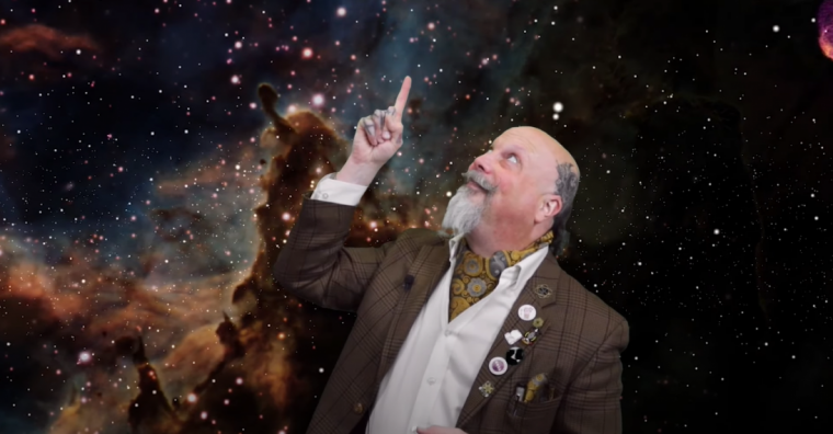 Orbax standing in front of a galaxy image looking up and pointing up at the sky.