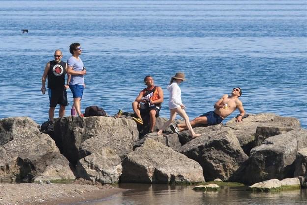 Image of young people lounging and socializing on rocks with water in background and beach in foreground