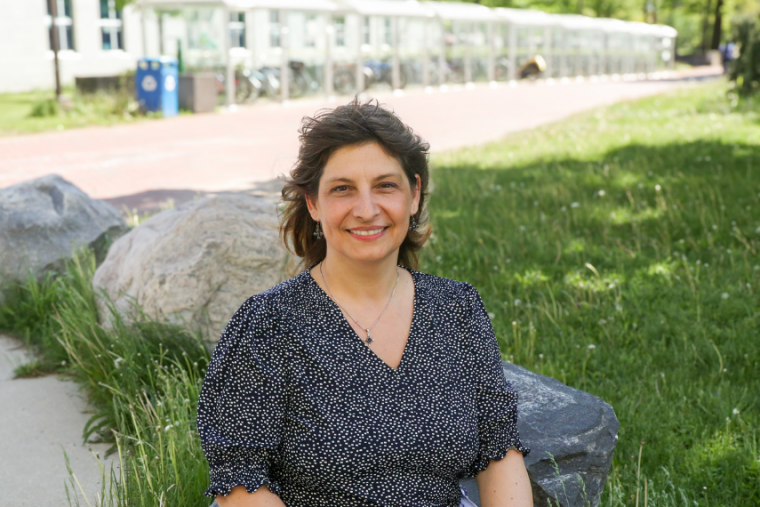 Dr. Monica Cojocaru sitting on a rock on campus in the summer, smiling at the camera.
