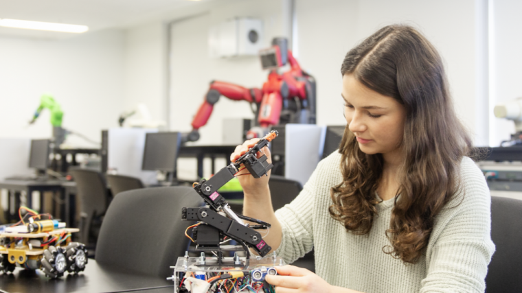 A student is seated in a lab surrounded by computers and robotic equipment. She is working on a wheeled robot with a robotic arm. Other robots and machinery are visible in the background, creating a modern robotics lab atmosphere.