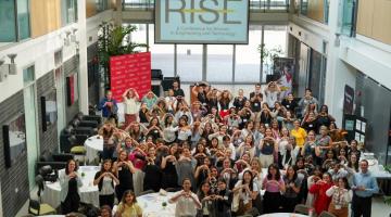 Large group of RISE attendees, speakers, and event organizers gathered, standing, in the Adams Atrium for an overhead group photo.