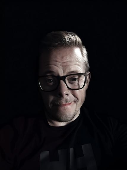 Dr. Daniel Gillis - a middle aged white man with dark rimmed glasses. He is lit from the right side, creating strong contrast with the dark background. 