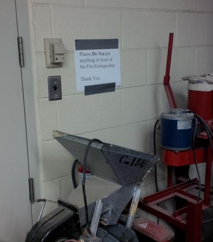 picture of a fire extinguisher blocked by equipment with a sign posted over it saying not to block the fire extinguisher