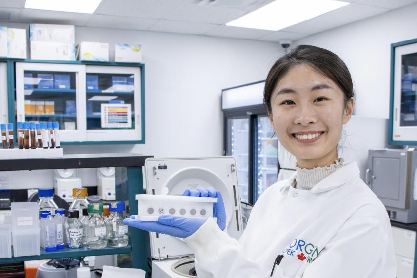 Maggie Xue wearing a labcoat and smiling for the camera. A lab can be seen in the background