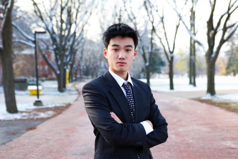 Thomas Deng, dressed in a suit, stands with his arms crossed, facing the camera confidently.