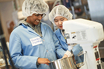 Two students in lab coats and hair nets working with a mixer in a lab.