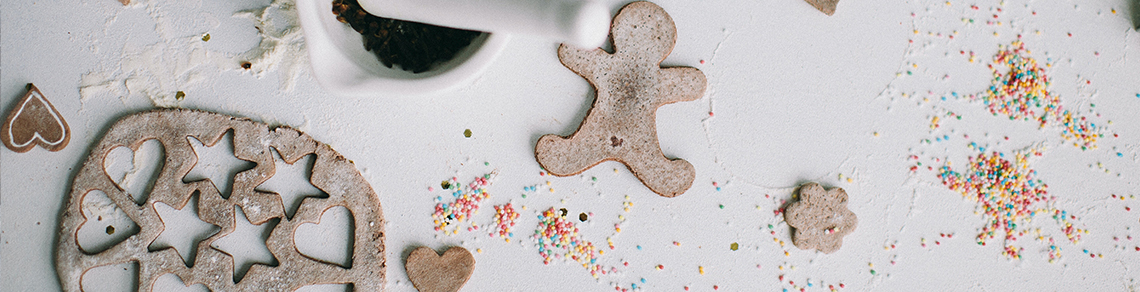 Image of gingerbread dough, with star and heart cookie shapes cut out of it and a gingerbread person cookie. There is flour and sprinkles on the surface of the counter alongside a mortar and pestle..