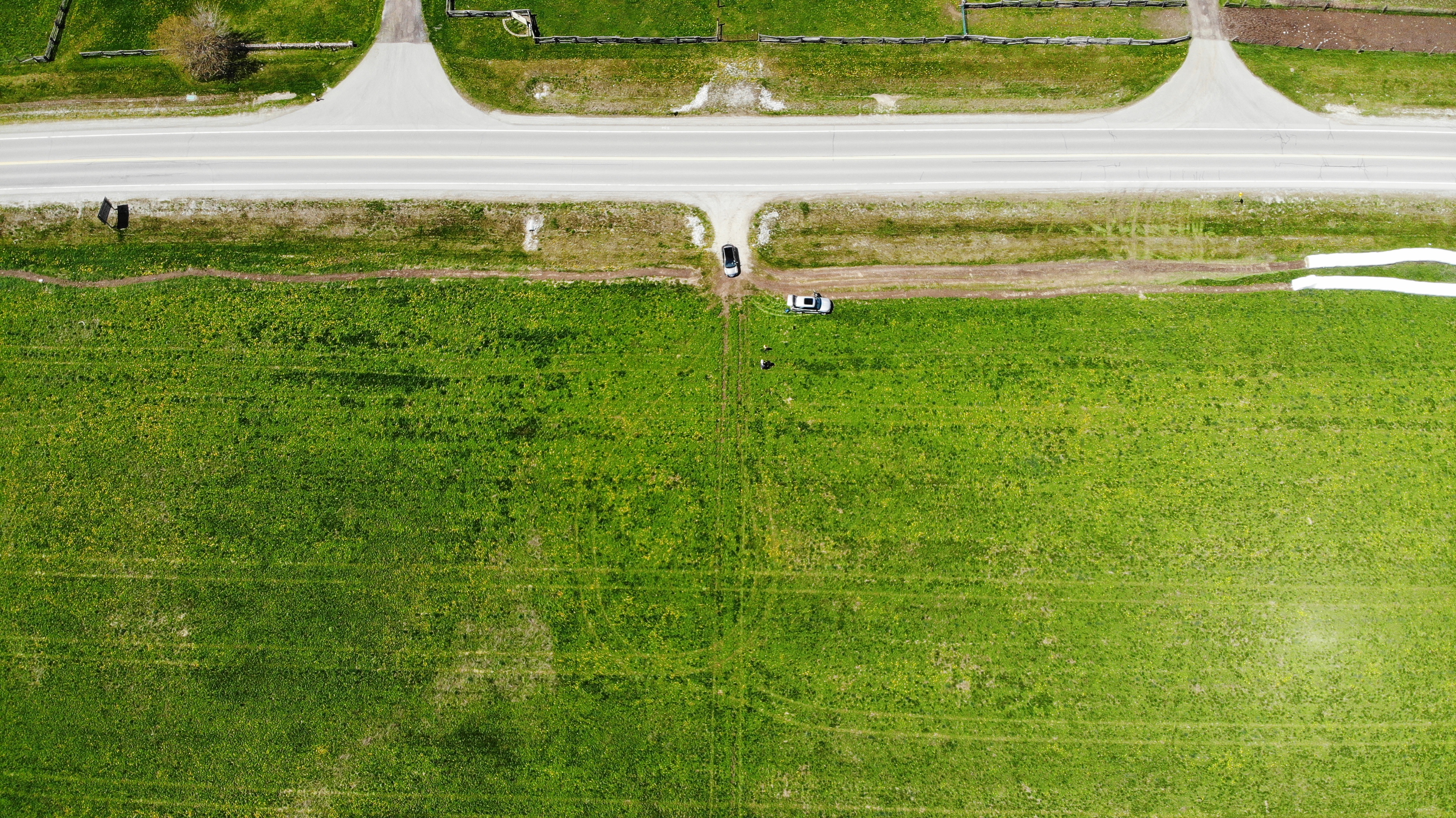 An aerial view of a green field with a road bordering and two cars turning off onto a dirt road.