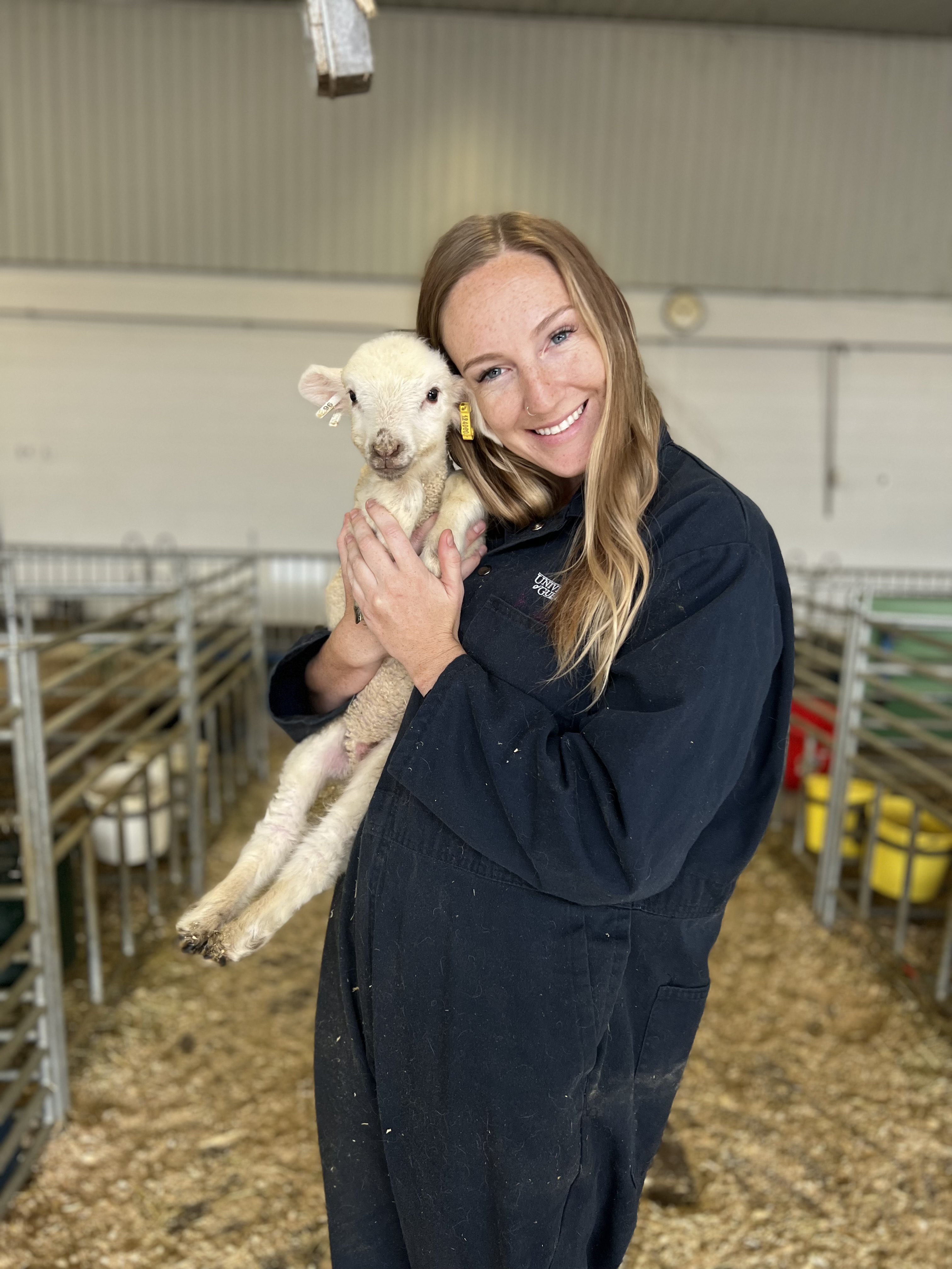 Researcher in coveralls smiling while cuddling a 10day old white lamb 