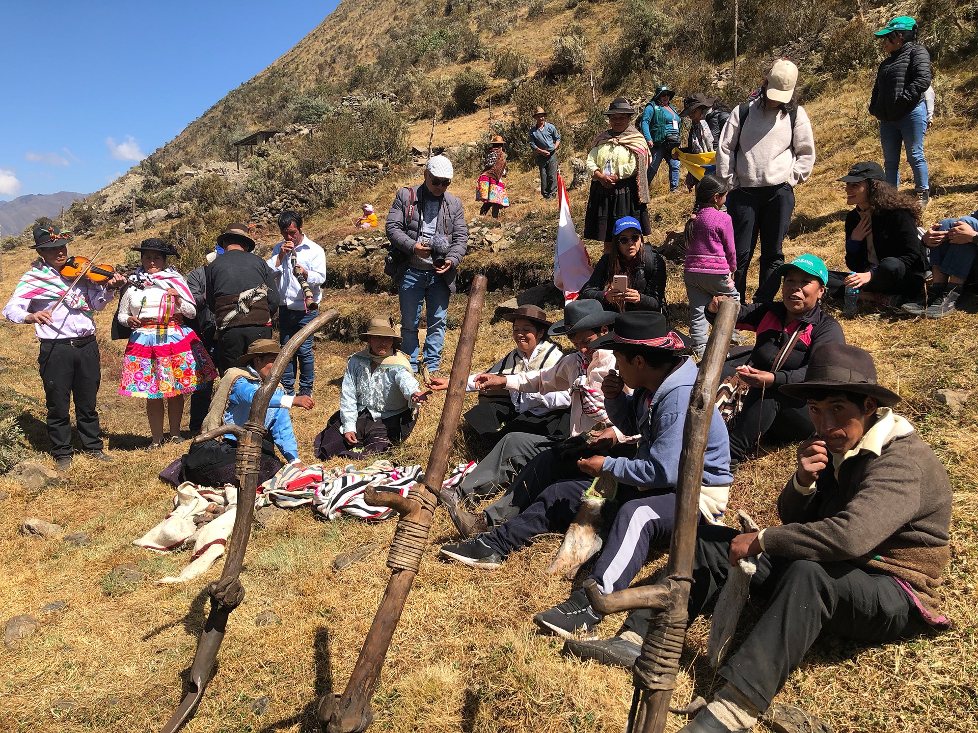 A group of people gathering on the side of the mountain. Some are sitting while others are standing. A few people have musical instruments including a horn,  tambourine and violin.
