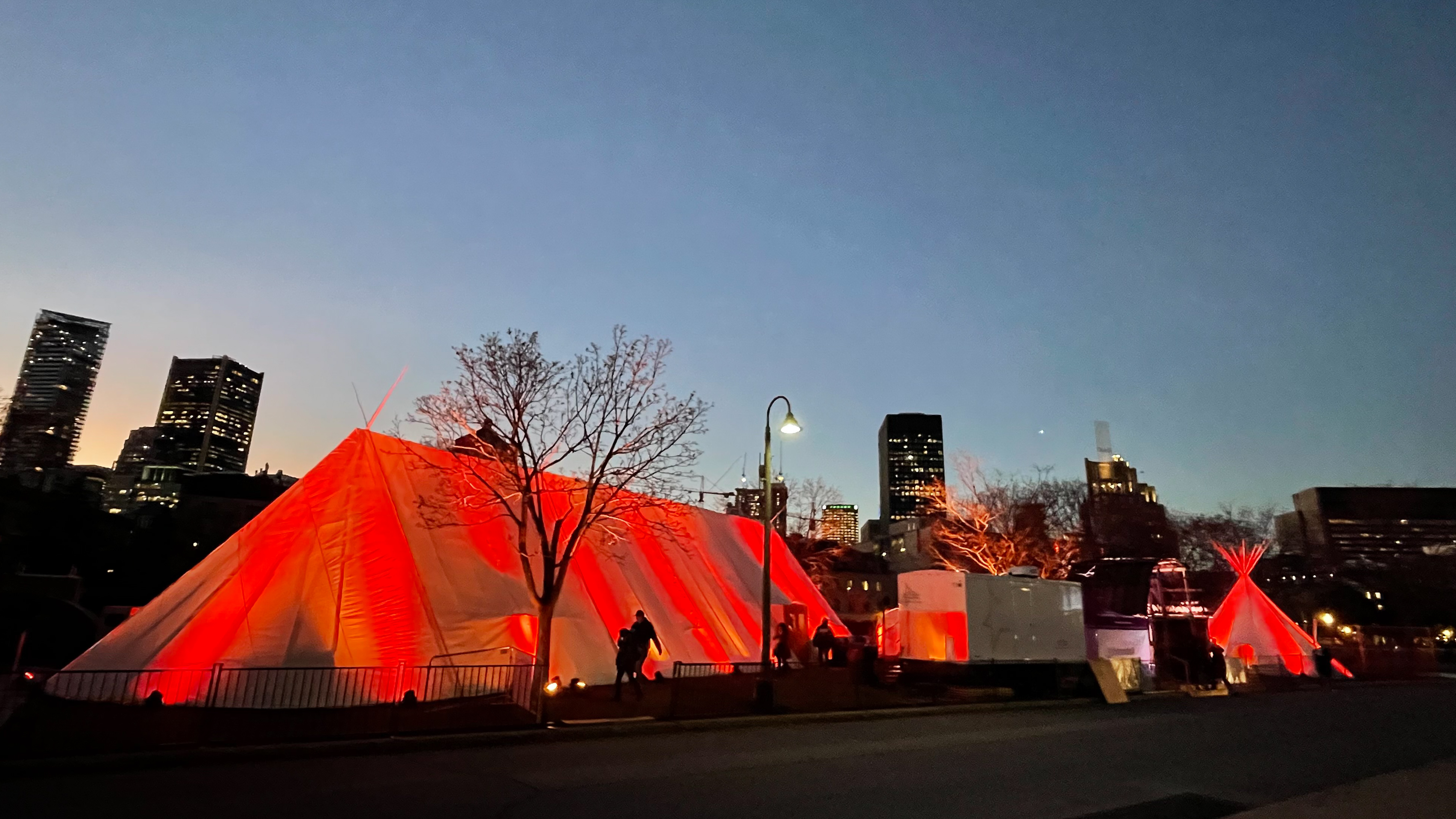 An Indigenous village in Montreal at night. There is a big tent and a teepee that are glowing red.