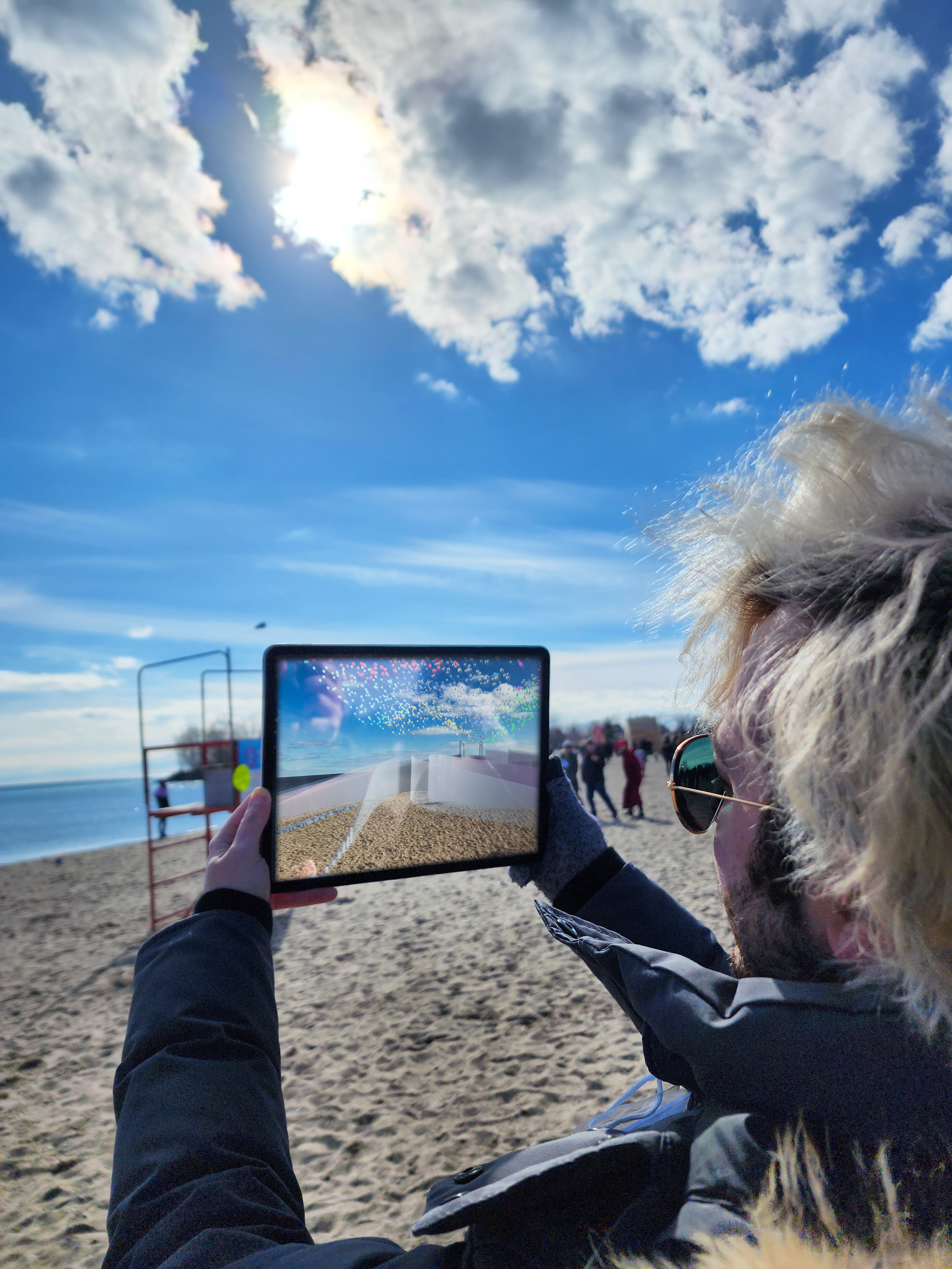 A person holding up a iPad on a beach with a big blue sky with white clouds. The scenery is reflected back at the person with the iPad.