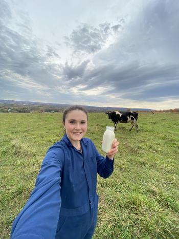 A woman in a field holding a jar of milk with a black and white cow behind her