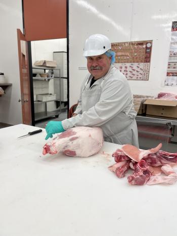 A man wearing a white lab coat, white helmet and blue gloves deboning a ham. Next to him are pieces of the pork.