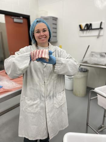 A woman in a white lab coat and a blue hair cover holding several slabs of bacon in her gloved hands.