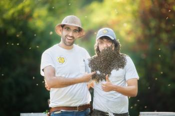 Two people standing together outside. One of the people has bees around his face in the shape of a beard.