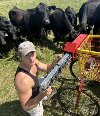 Man kneels in pasture surrounded by black cows as he works on a machine attached to a red and yellow cart.