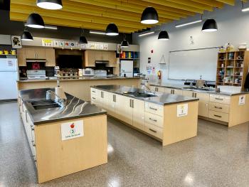 A modern kitchen classroom, with counters that are at an appropriate height to be accessible for 6-12 year olds to learn how to cook and follow along with their teacher. 
