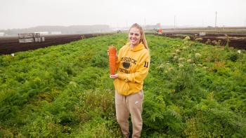 A woman standing in a field of crops with a giant carrot in her hands.