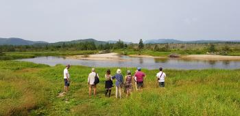 A group of eight people standing in tall grass in front of a river listening to the sounds of nature