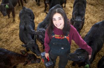 A dark-haired woman wearing a purple sweatshirt and black overalls standing with a several black bull calves