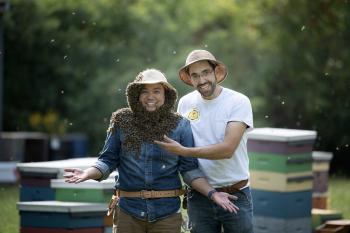 Two beekeepers standing side by side. One beekeeper's face and neck are covered in bees.