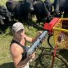 Man kneels in pasture surrounded by black cows as he works on a machine attached to a red and yellow cart.